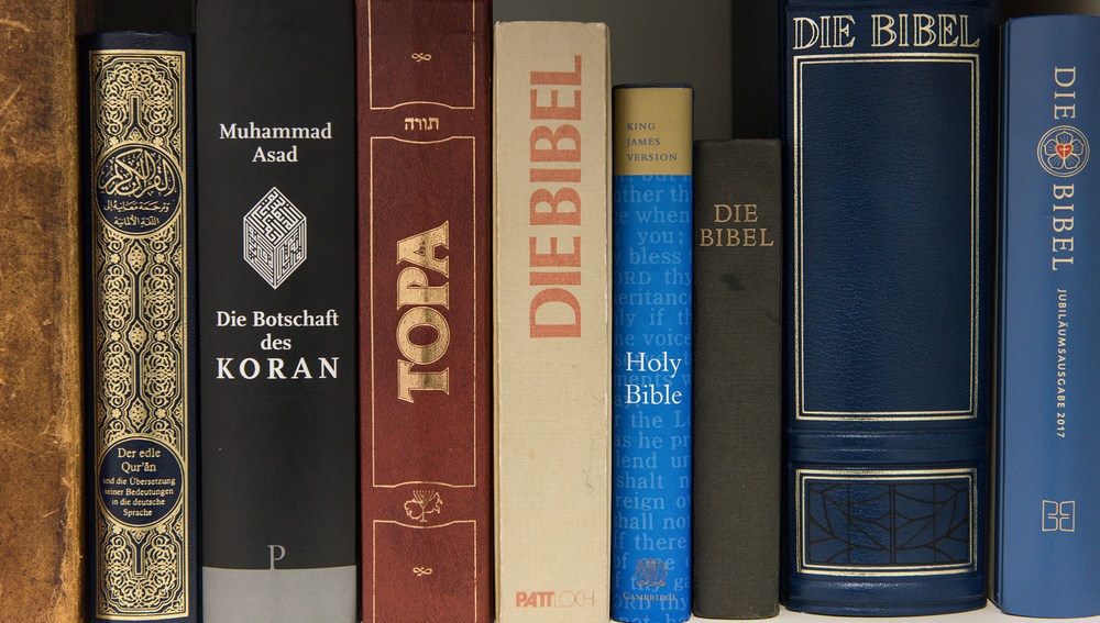Blick auf die Buchrücken verschiedener Ausgaben des Koran, der Thora und der Bibel nebeneinander in einem Bücherregal. Die Bücher sind die heiligen Schriften der drei abrahamitischen Religionen Islam, Judentum und Christentum.